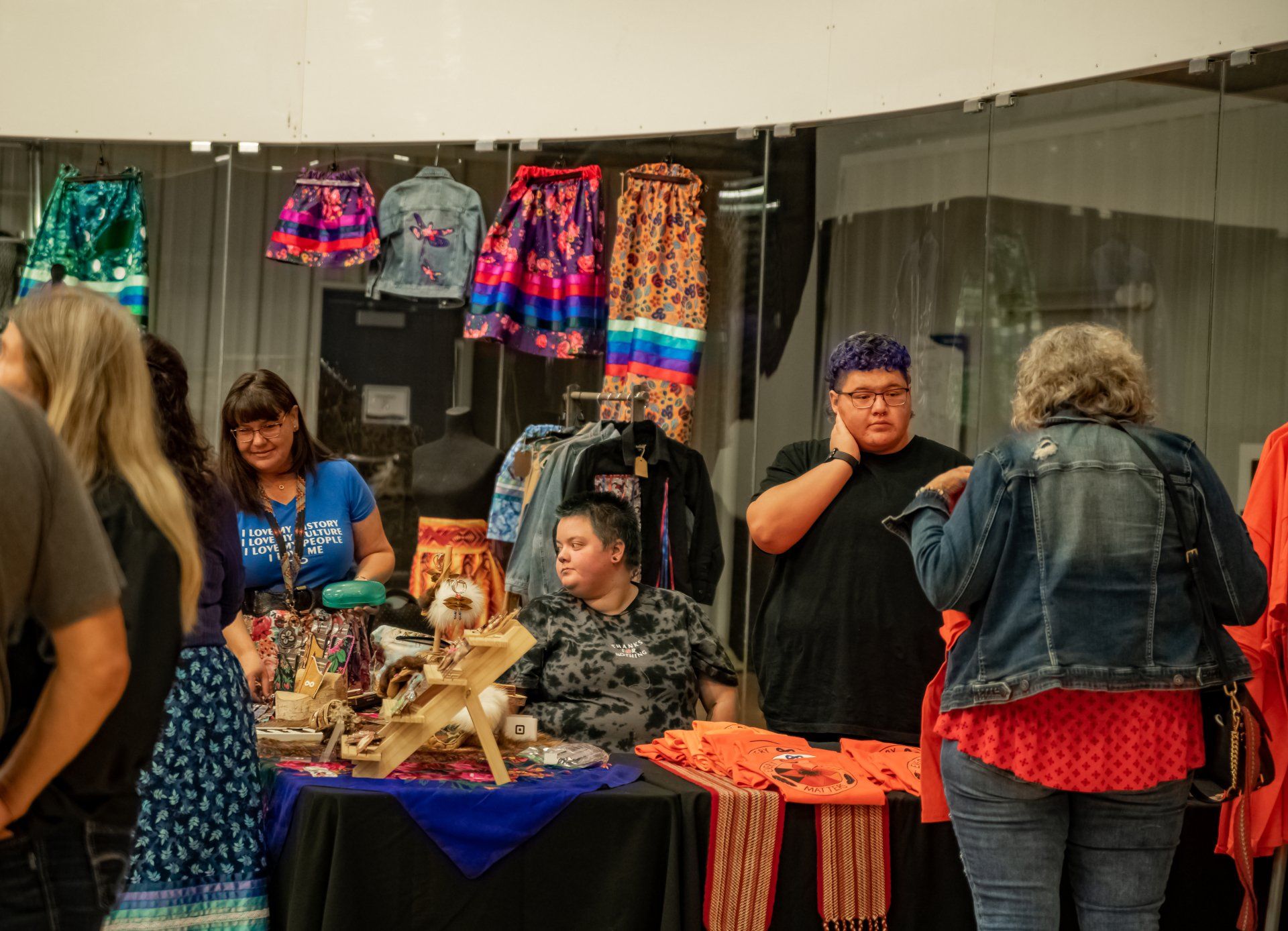 People at a craft fair, browsing and buying handmade clothing and jewelry at a booth with colorful skirts and beaded items.