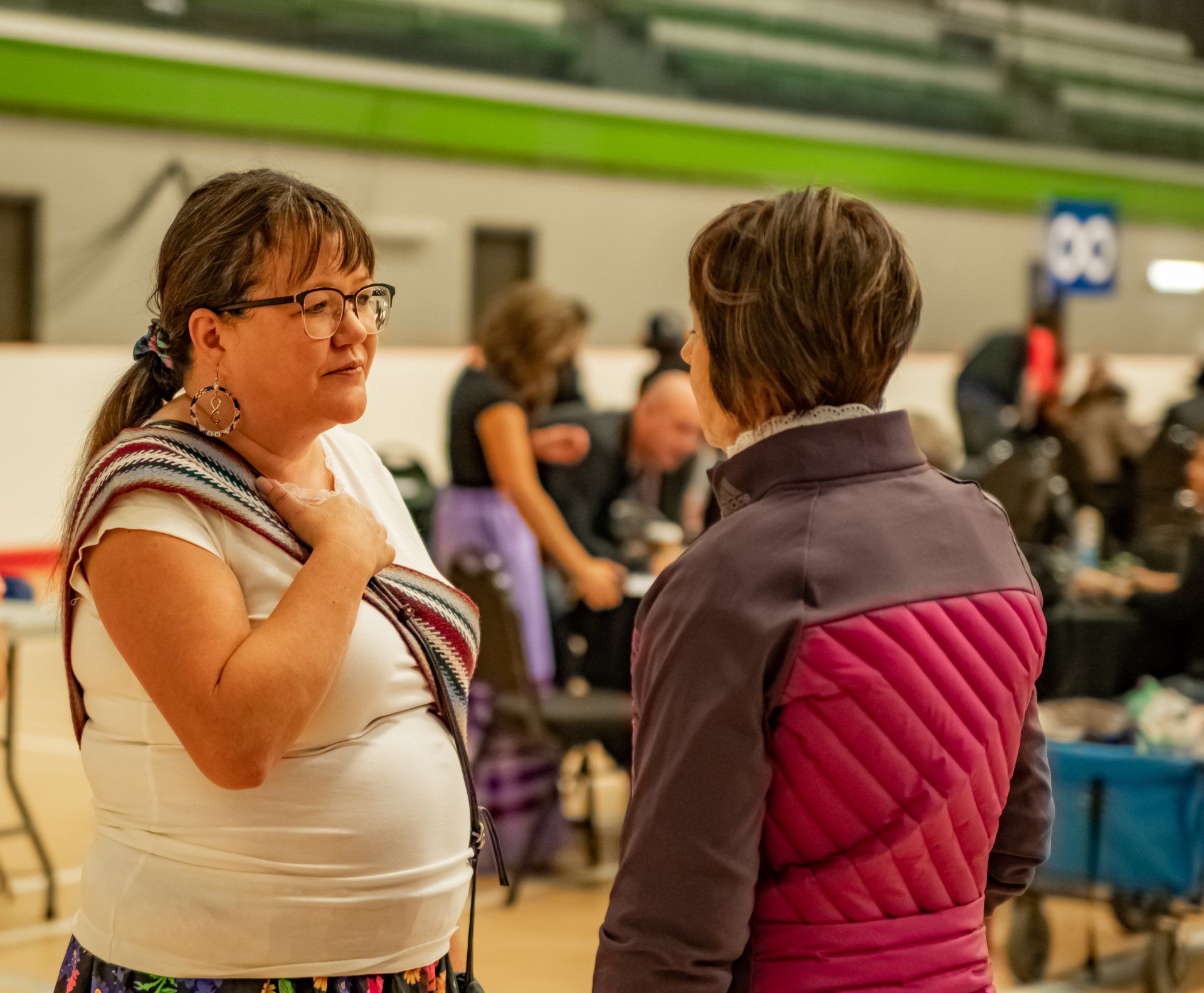 Two women talking in a gym. One wears glasses, white shirt, patterned wrap; the other, a purple jacket. Background: people, tables.