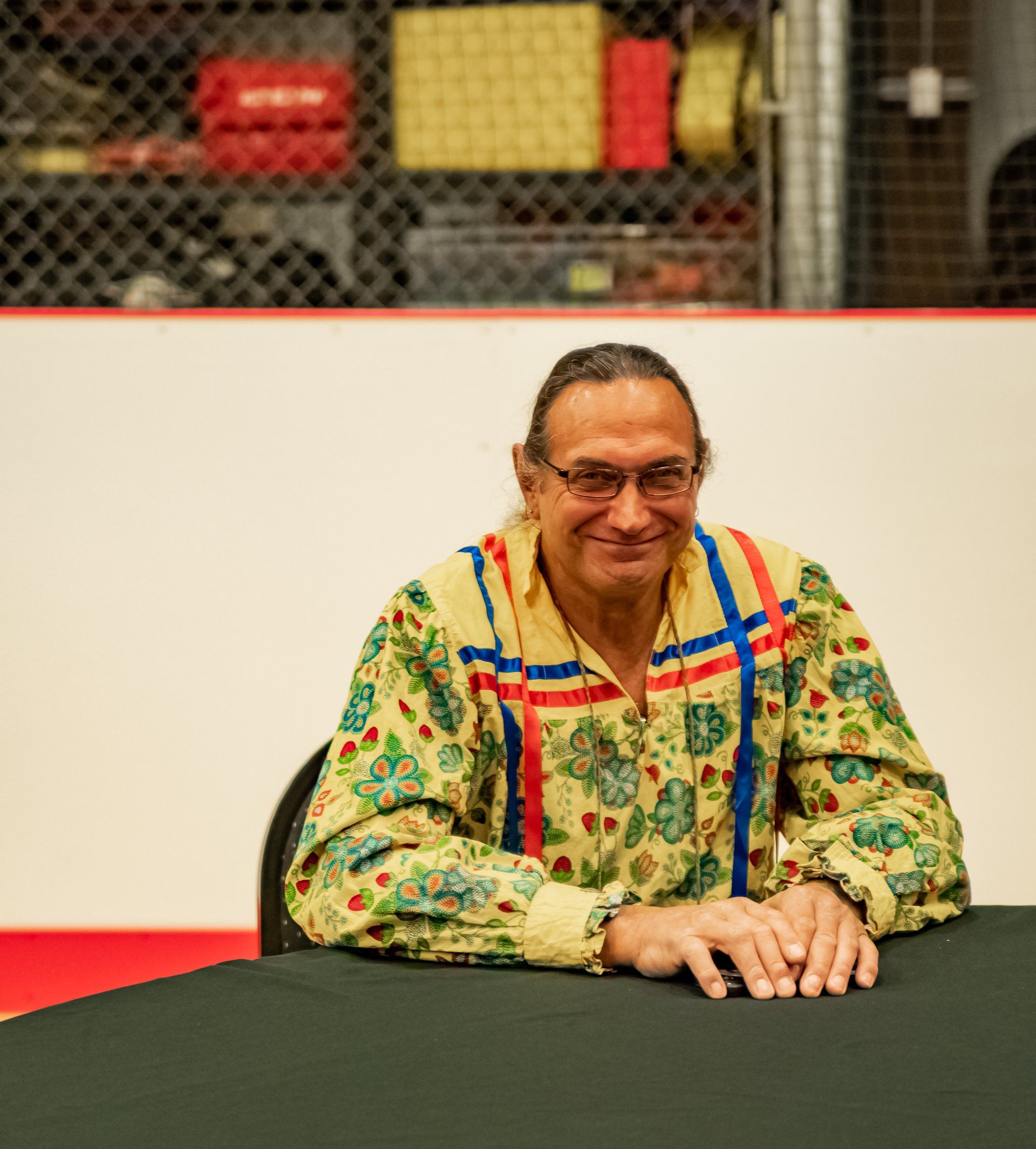 Man in a patterned shirt sits at a table, smiling. Red and blue accents on his shirt. Indoor setting.