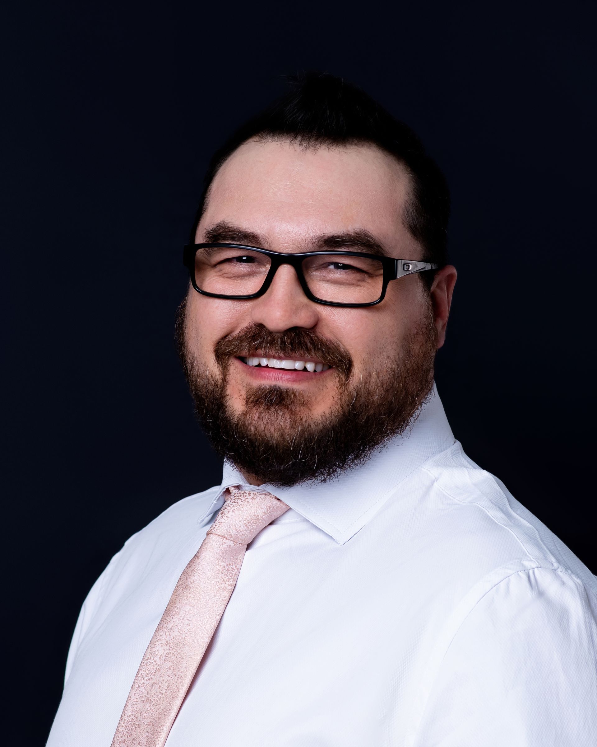 Man with glasses and beard, wearing a light pink tie and white shirt, smiling against a dark background.