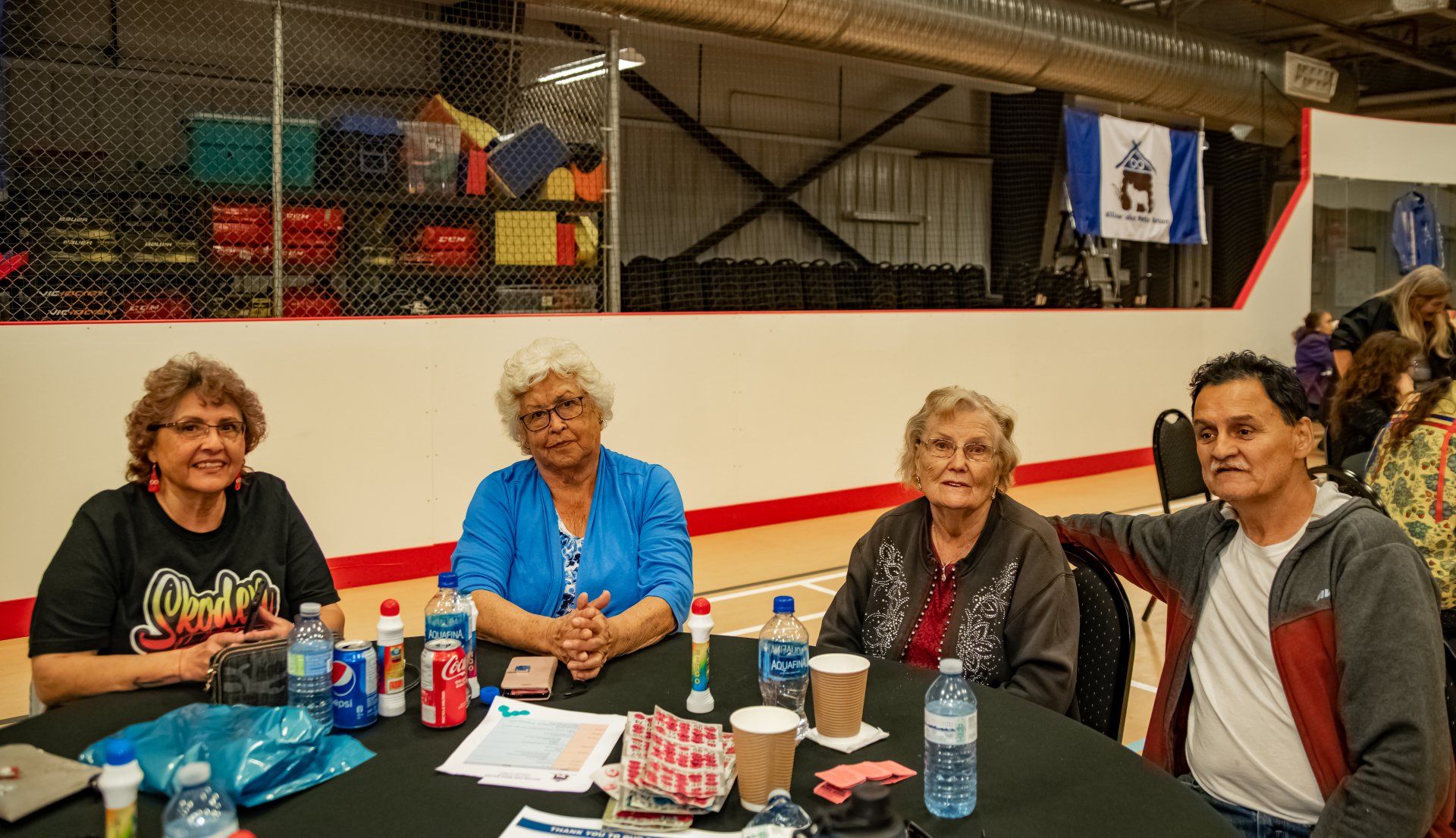 Four people at a table in a gymnasium. They are smiling, with drinks and supplies on the table.
