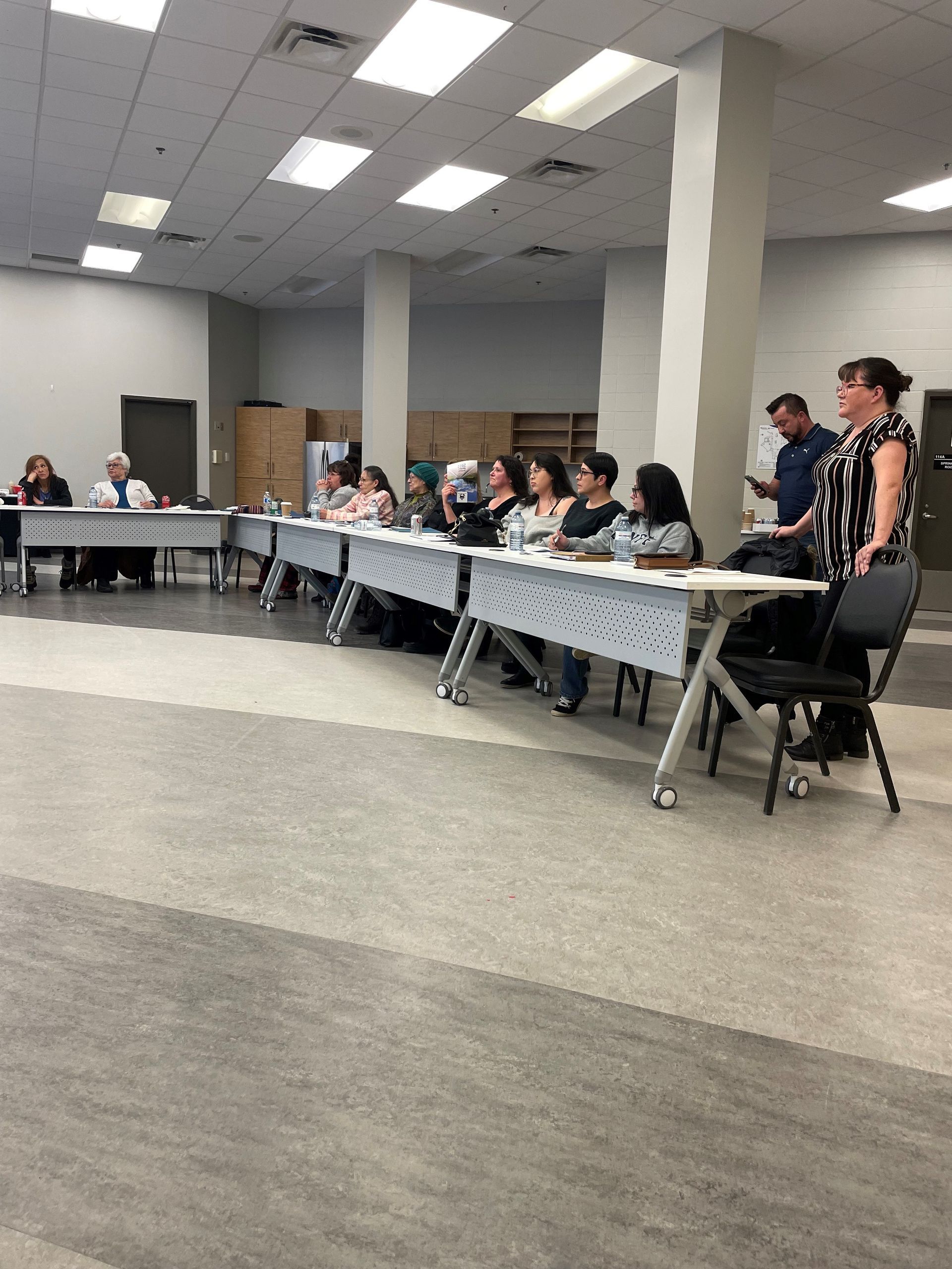 People seated at tables in a room, appearing to be a meeting. White walls, gray floor.