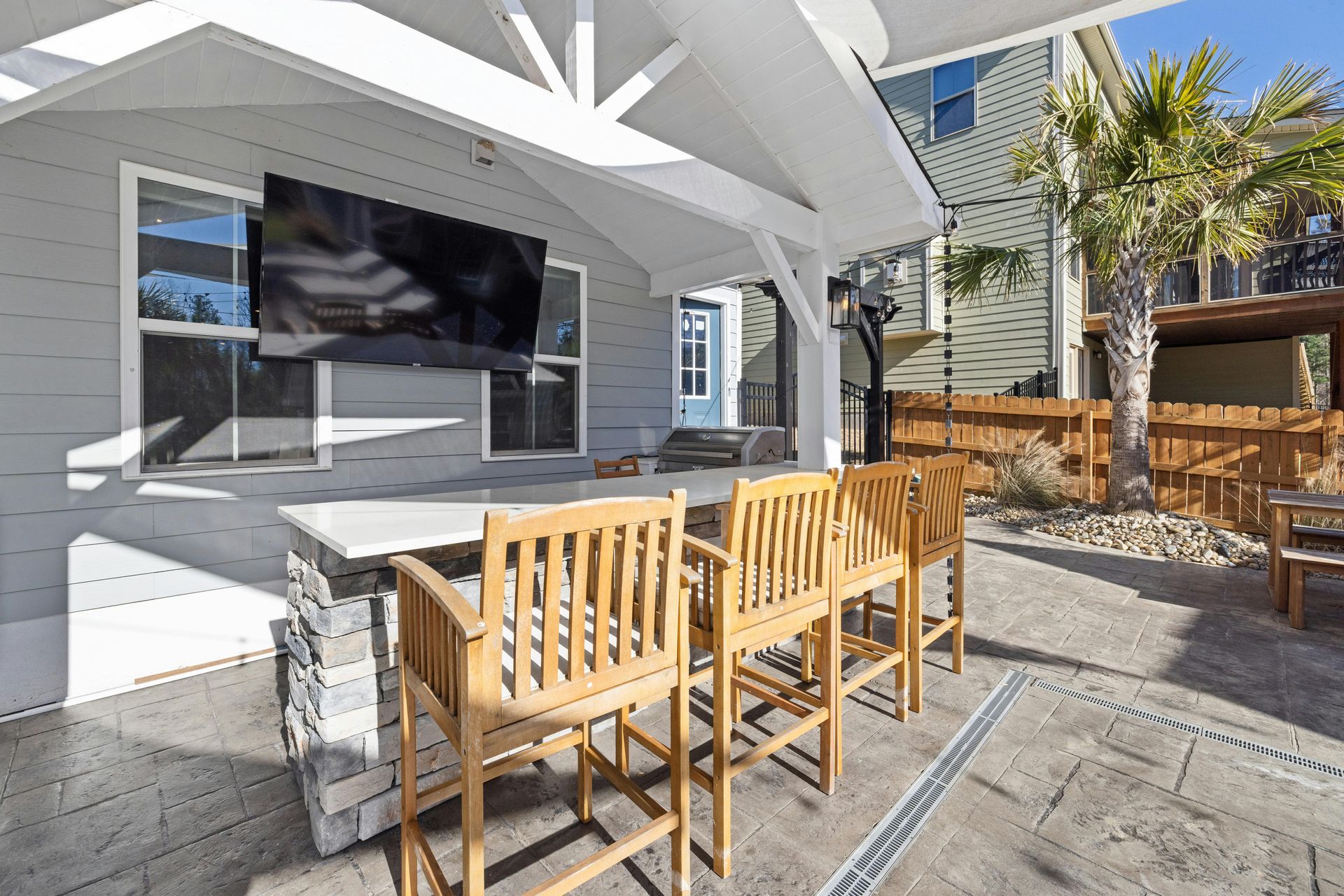 Outdoor patio with wooden chairs and tables under a white pergola beside a gray house