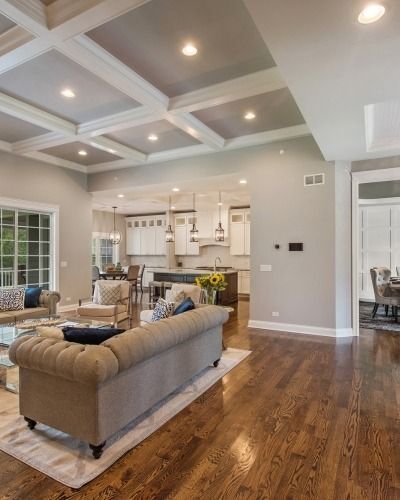 Spacious living room with coffered ceiling, hardwood floors, tan sofa, and view into a bright kitchen.