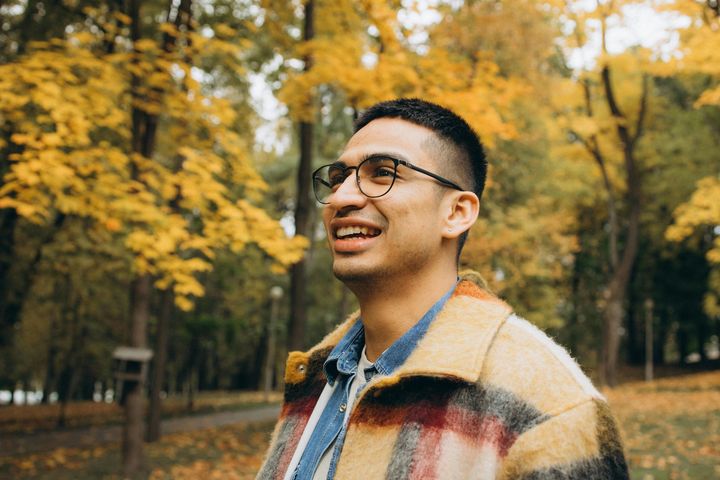 Smiling person in glasses wearing a plaid jacket, standing in a tree-lined park with autumn leaves