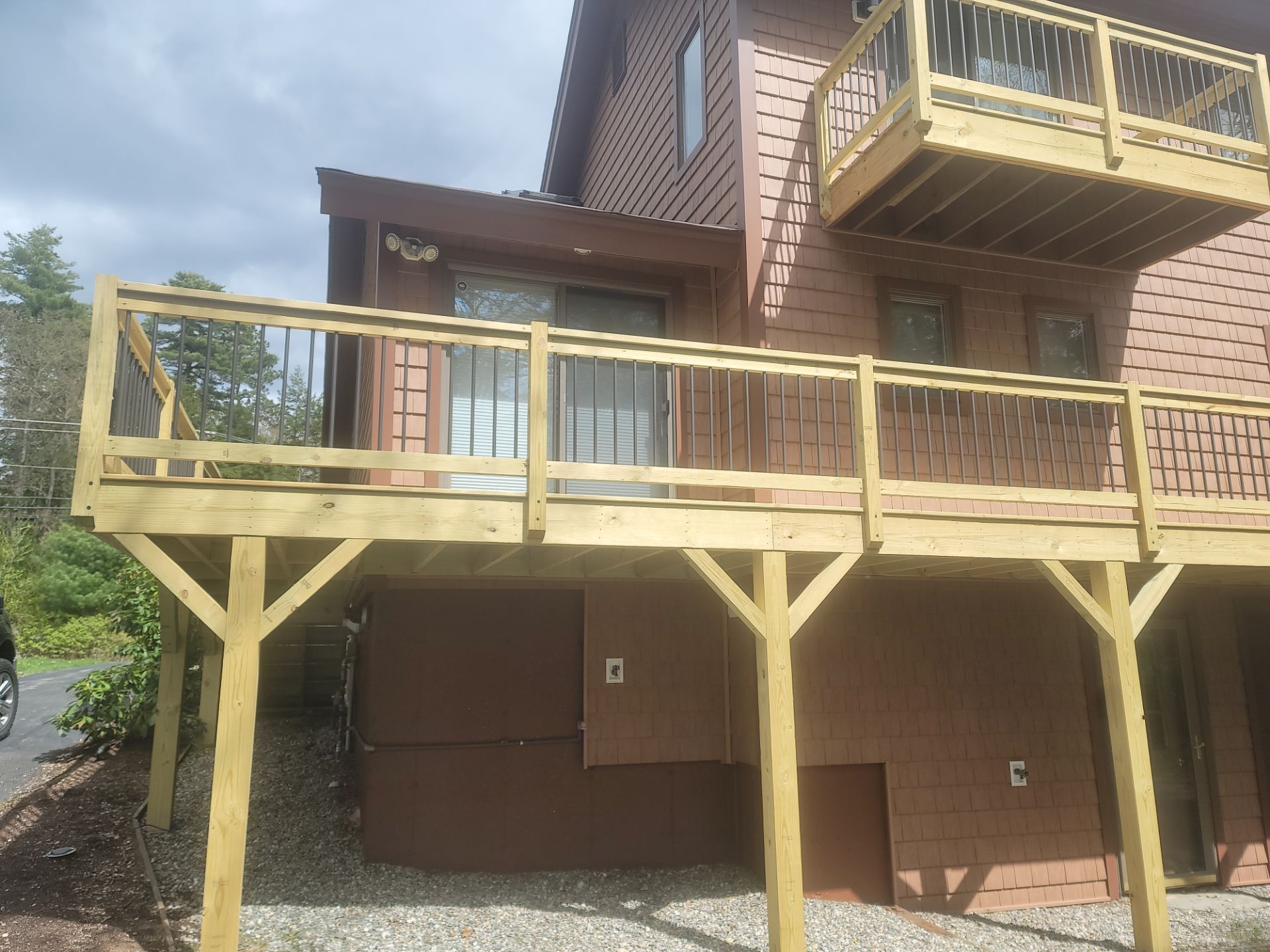 Wooden deck and railing attached to a brown house, with support posts over a shaded ground area.