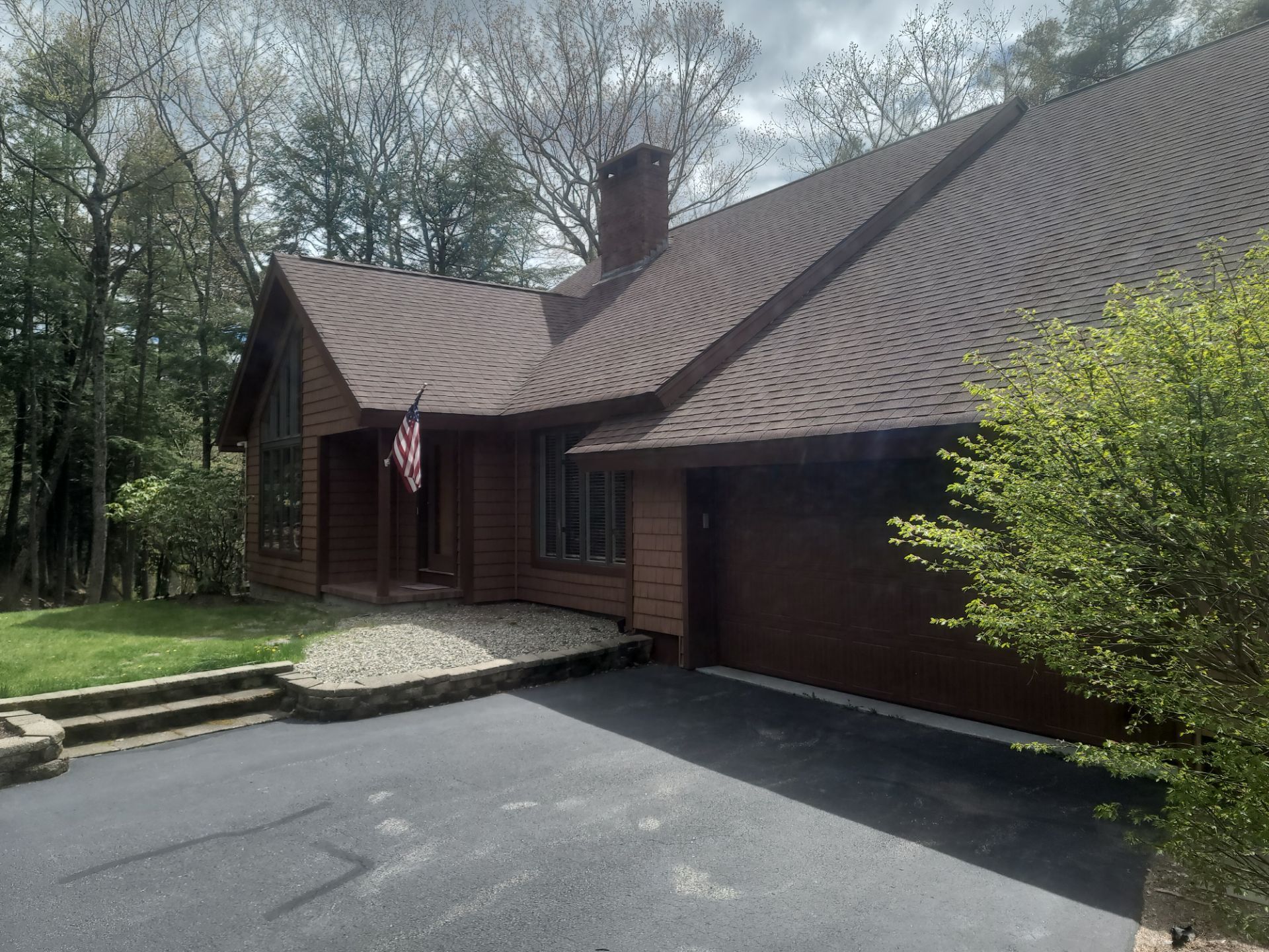 Wooden house with a steep shingled roof, driveway, and American flag beside a wooded yard