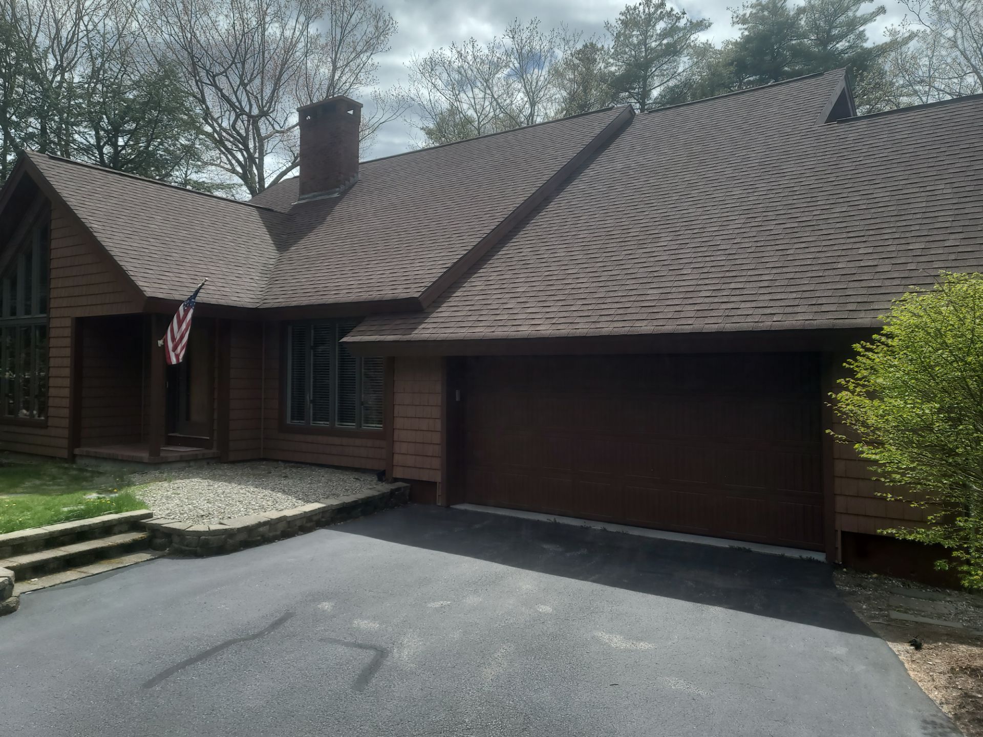 Suburban brick house with dark shingle roof, attached garage, and driveway under cloudy sky