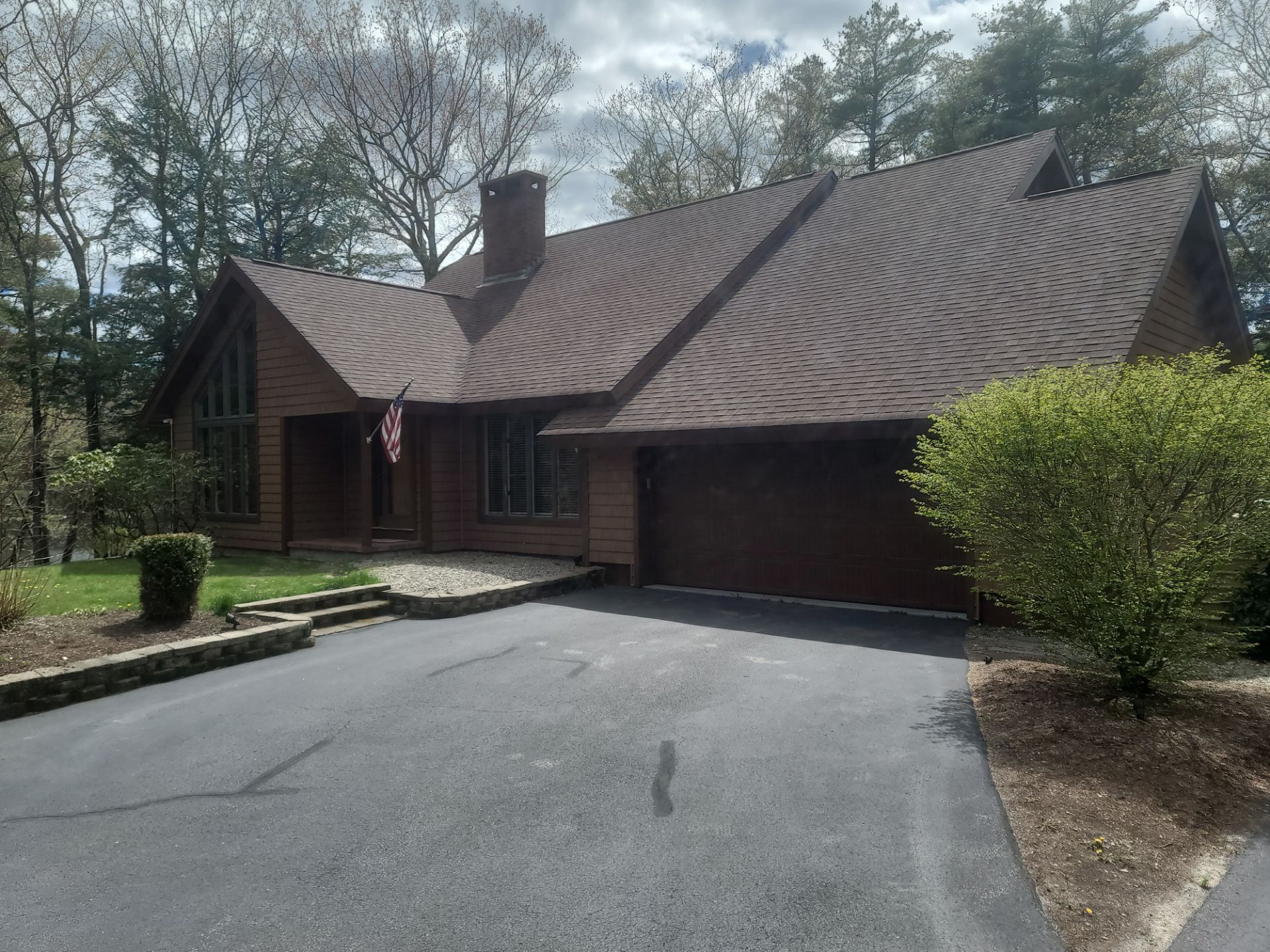 House with brown roof and garage at end of a curved driveway, surrounded by trees and shrubs