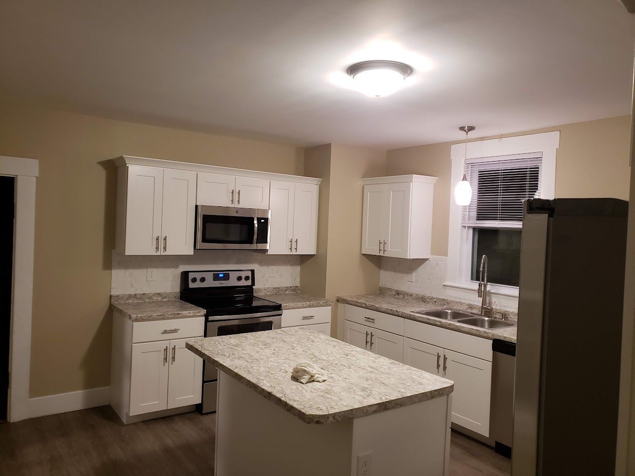 Small beige kitchen with white cabinets, granite island, stove, dishwasher, and window over the sink.