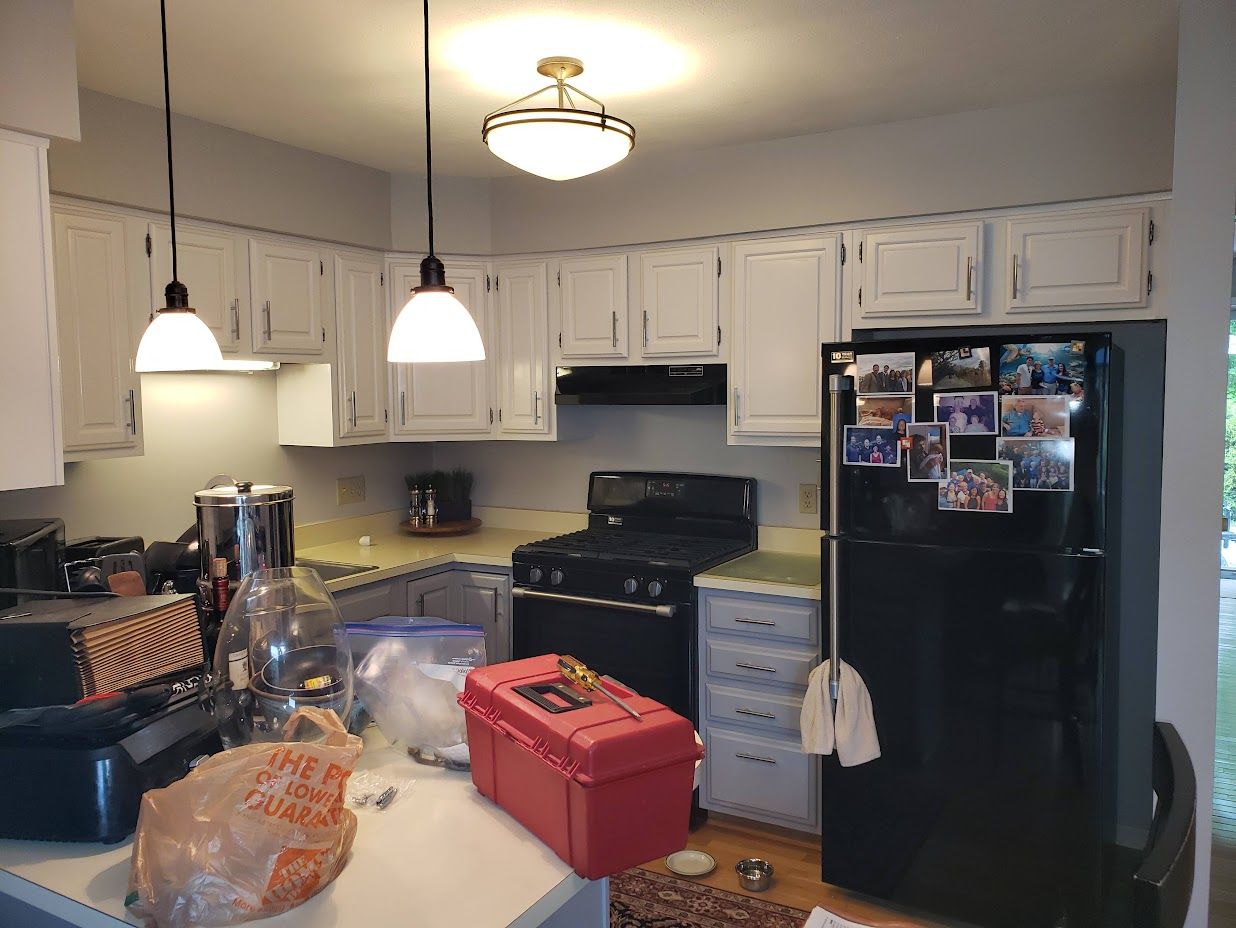 Cluttered kitchen with white cabinets, black stove and fridge, hanging lights, and a cluttered counter and island.