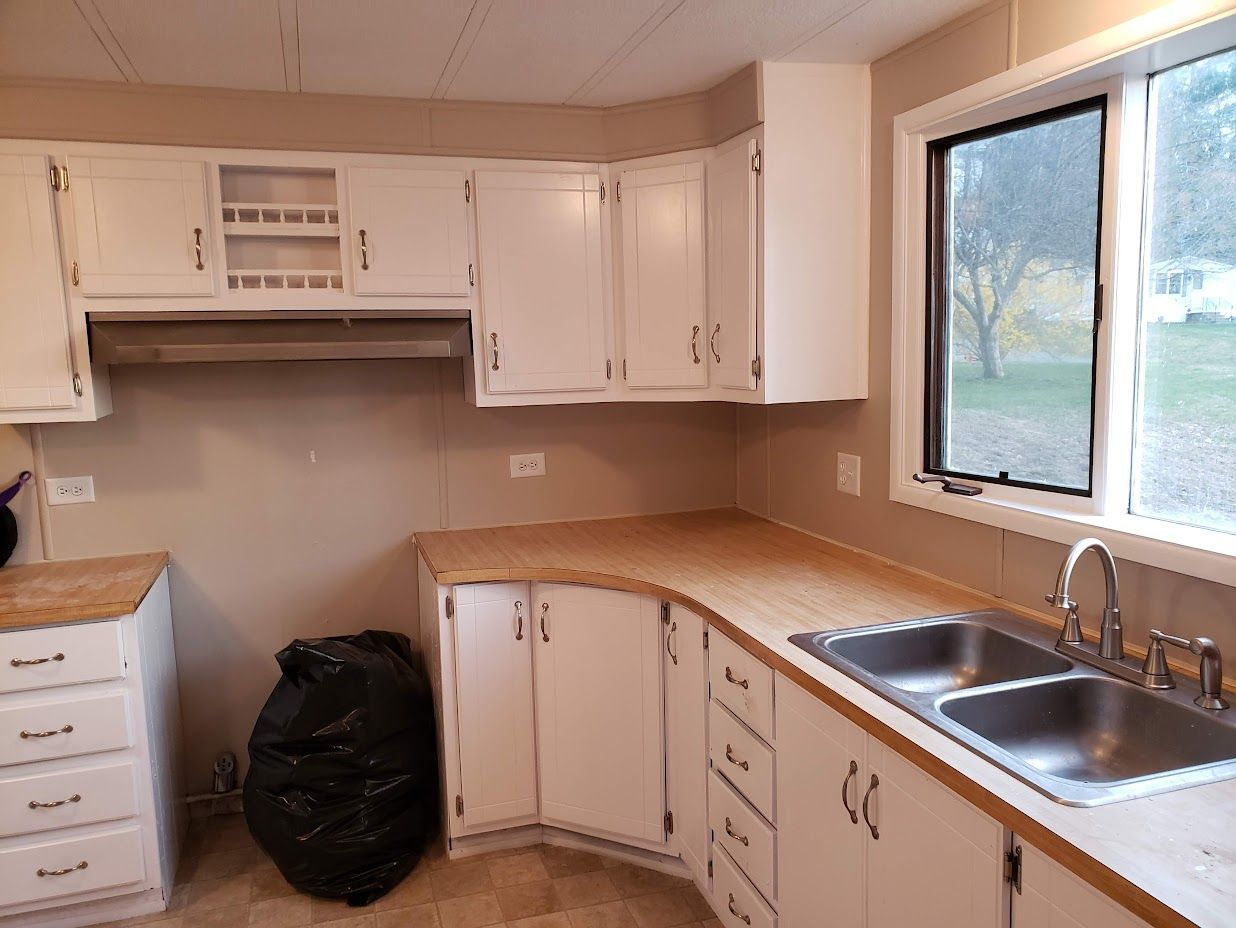 Empty kitchen with white cabinets, tan counters, and a double sink by a window.