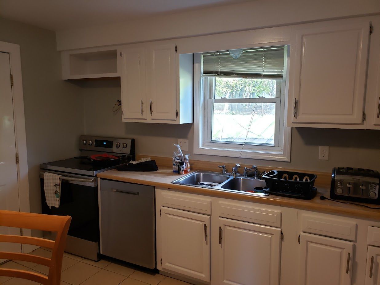 Small kitchen with white cabinets, stainless dishwasher, stove, sink under window, and wooden chair in foreground