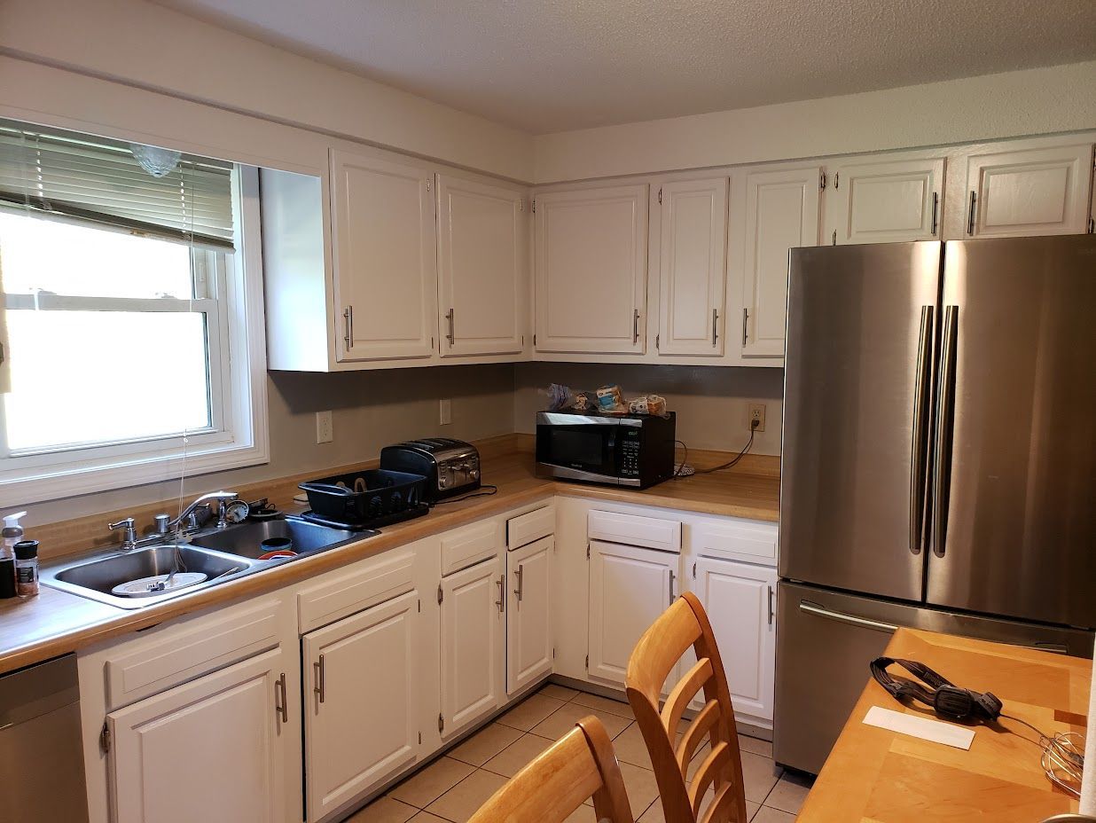 Bright kitchen with white cabinets, stainless fridge, wooden table, and window over the sink