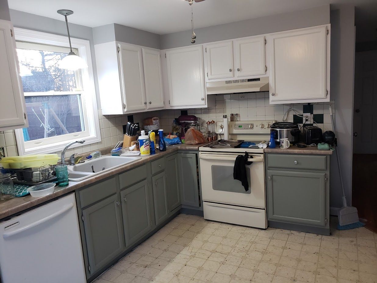 Small kitchen with white cabinets, gray lower cabinets, a stove, and a cluttered countertop by a window.