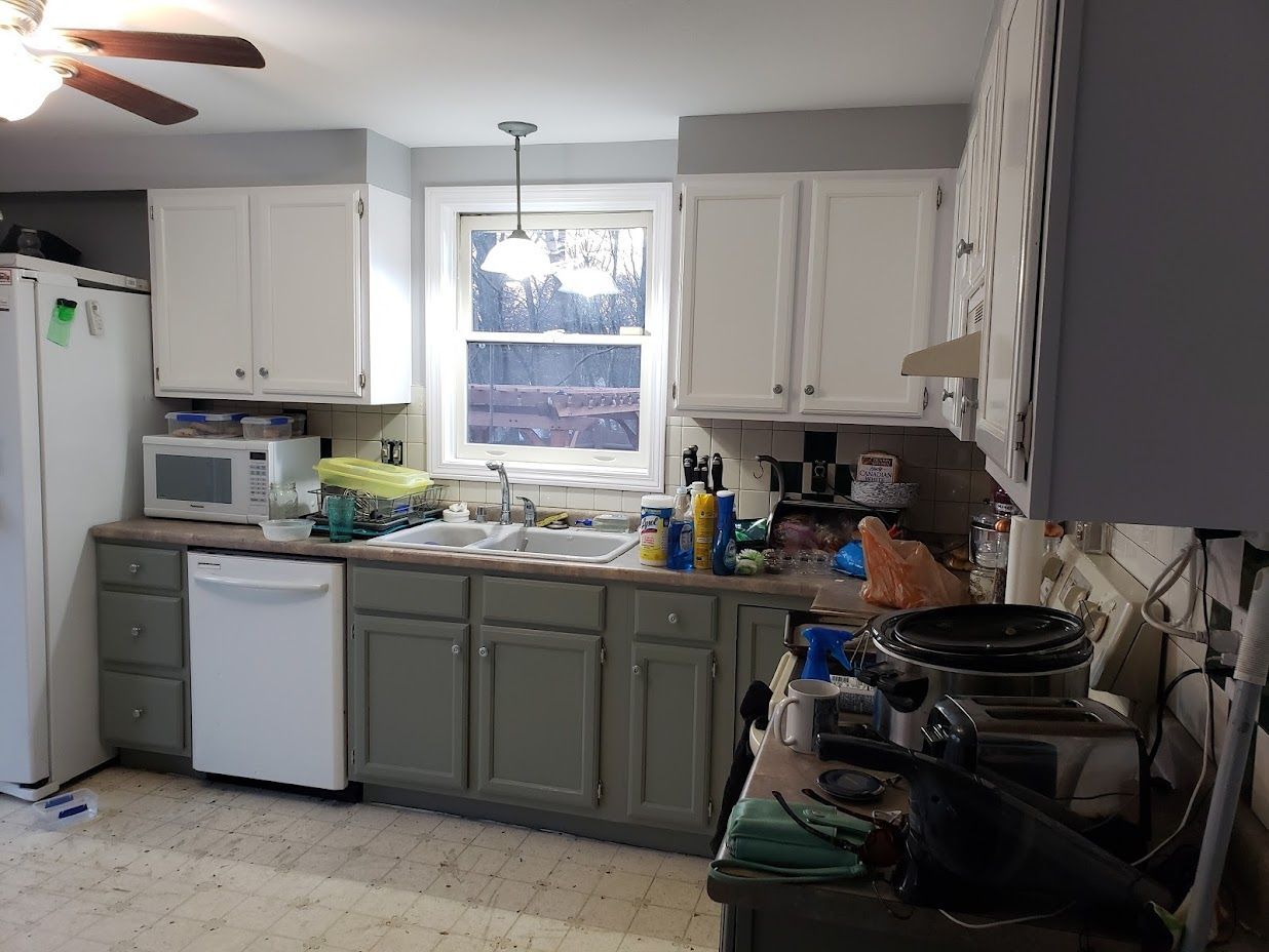 Small kitchen with white and sage cabinets, window above sink, and cluttered counters.