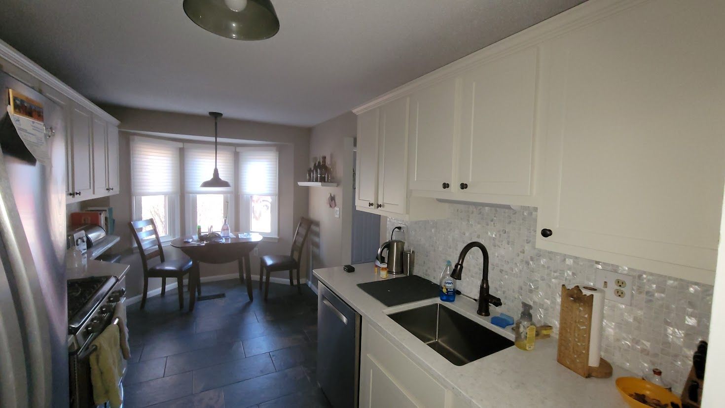 Long, narrow kitchen with white cabinets, black countertops, and a dining area by a bright window.
