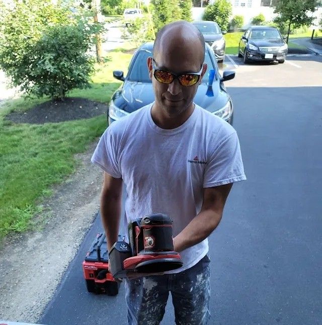 Man holding a red and black sander outdoors beside a parked car on a driveway