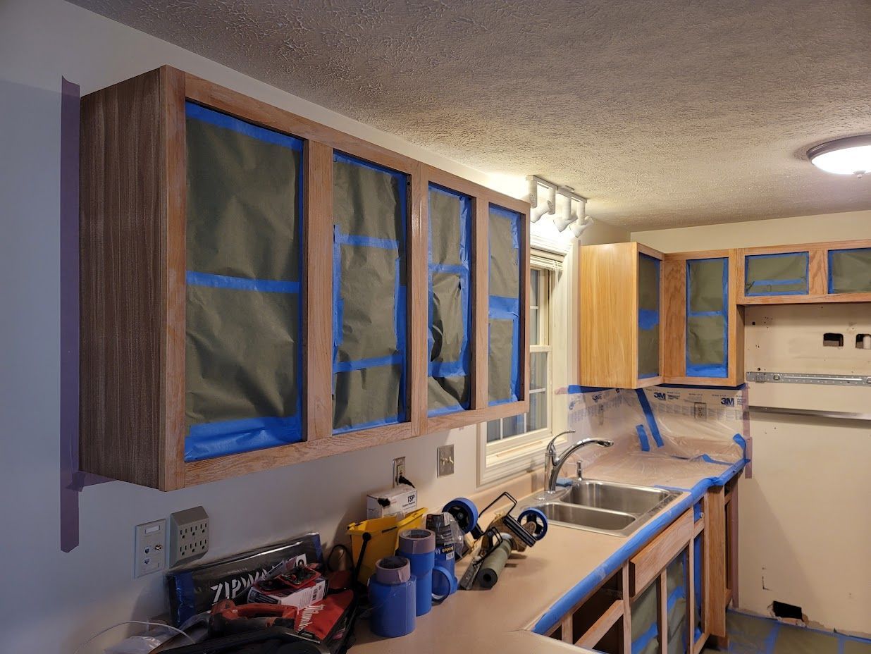 Kitchen under renovation with cabinets taped for painting and countertops covered with tools.
