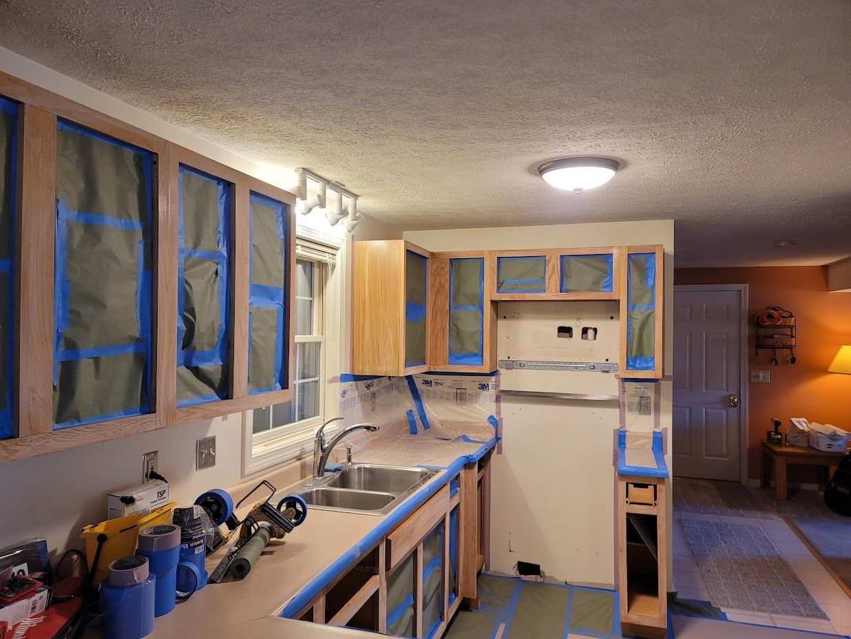 Kitchen under renovation with taped cabinets, covered windows, and a refrigerator in a compact workspace