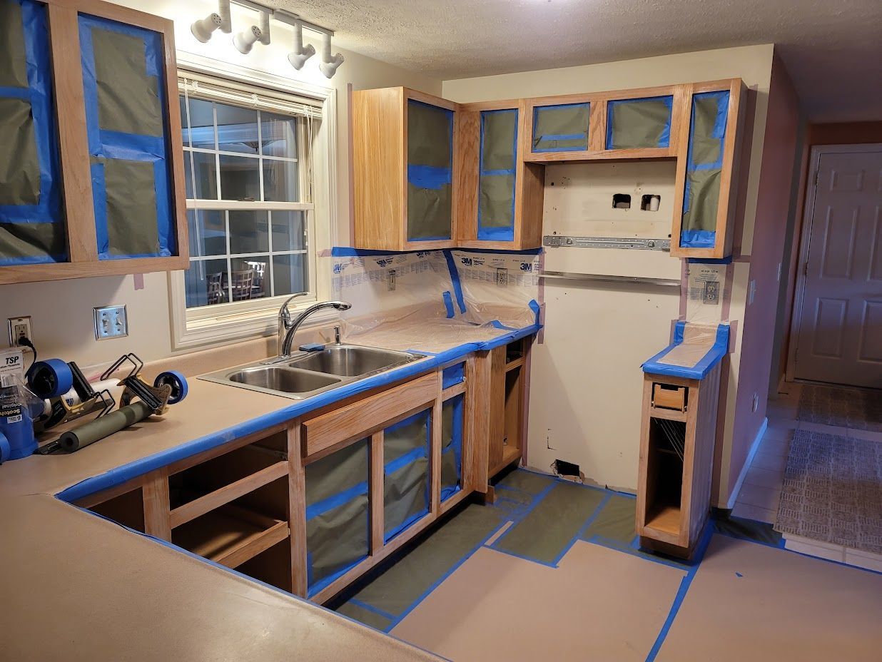 Unfinished kitchen with blue painter’s tape and paper masking cabinets, counters, and floor for painting.