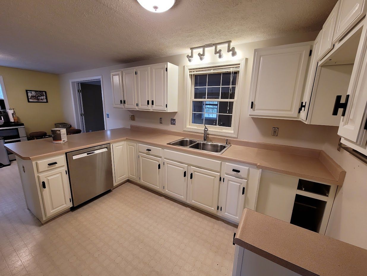 Bright kitchen with white cabinets, beige counters, stainless dishwasher, and a window over the sink.