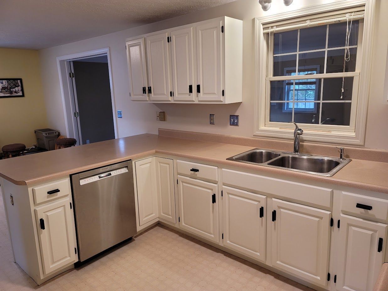 Bright kitchen with white cabinets, beige countertops, stainless dishwasher, and a double sink by a window.