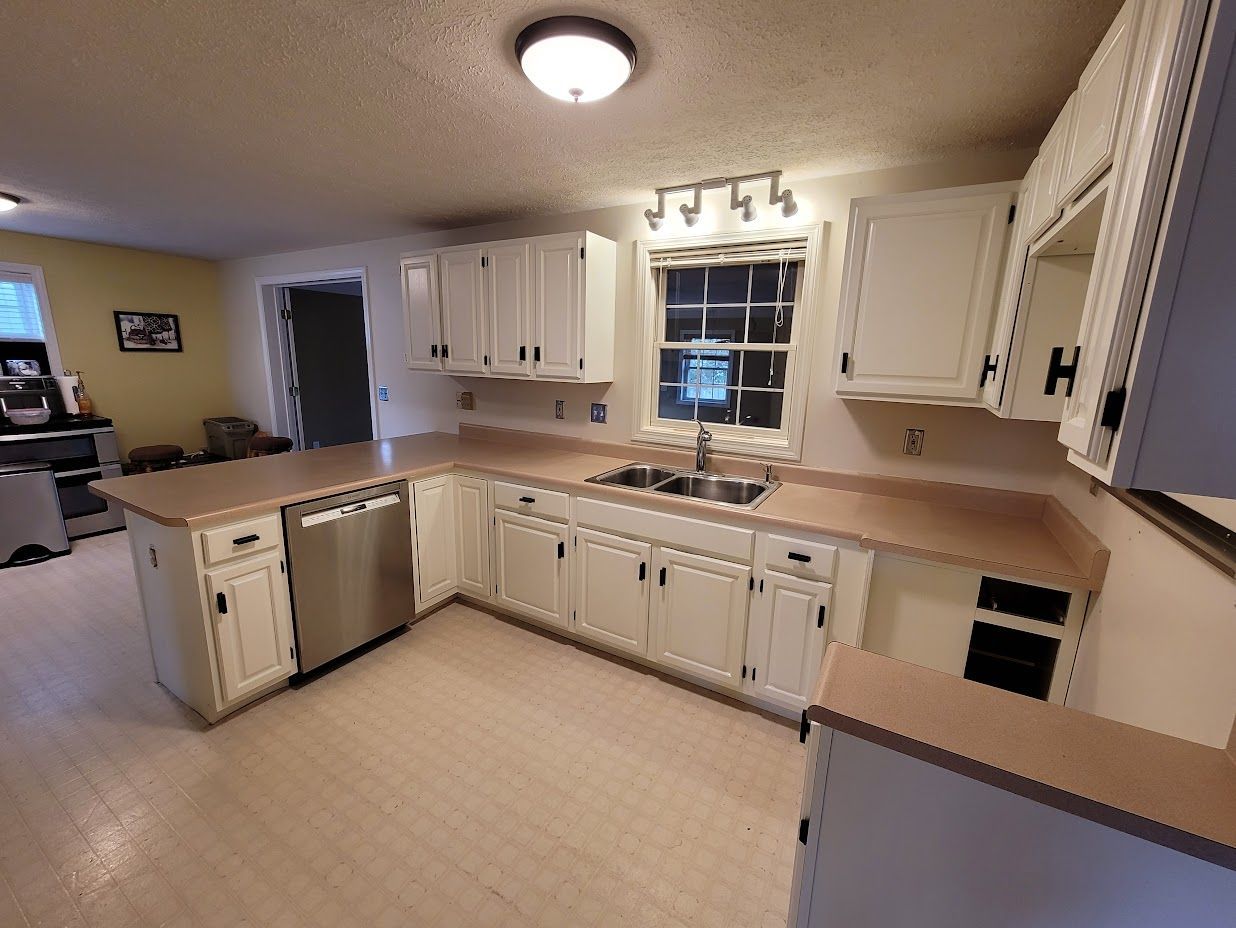 Bright kitchen with white cabinets, beige counters, stainless dishwasher, and a window over the sink.