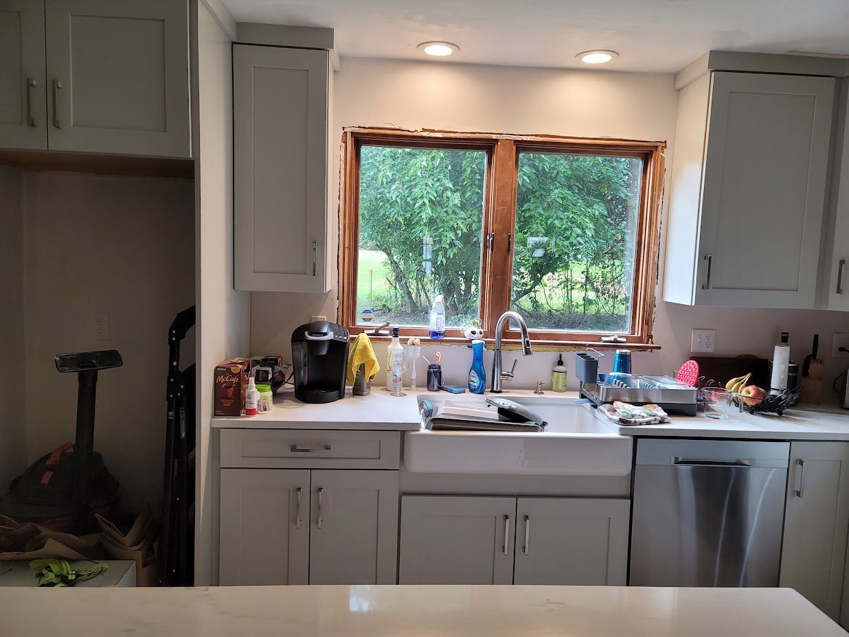 Kitchen sink beneath a window, with light cabinets, countertop items, and greenery outside.