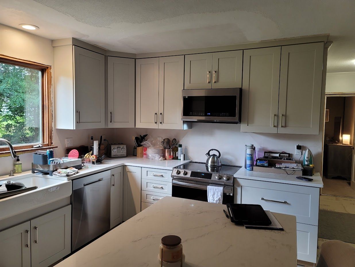 Bright kitchen with gray cabinets, stainless stove and dishwasher, large window, and an island in front.