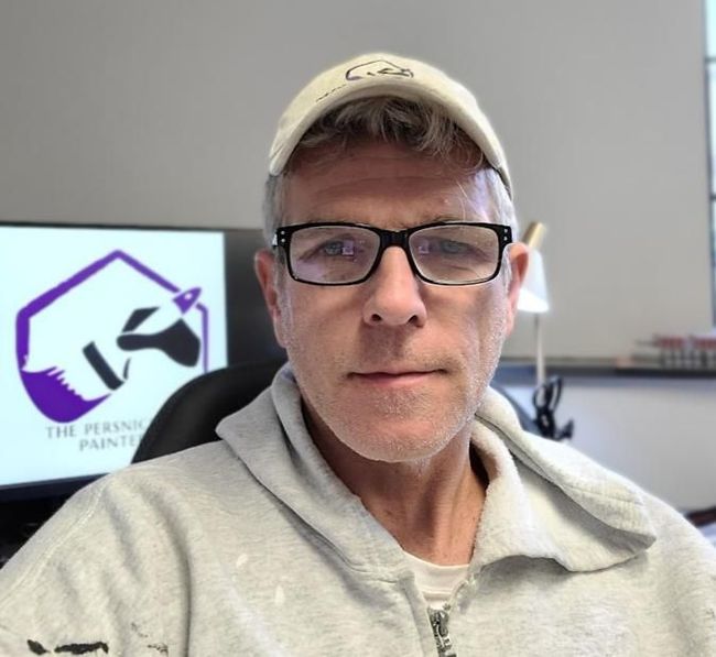 Man in glasses and cap seated indoors, with a computer monitor displaying a purple logo in the background
