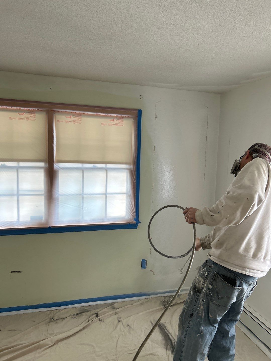 Person painting a room interior with blue tape around a window and paint supplies on the floor