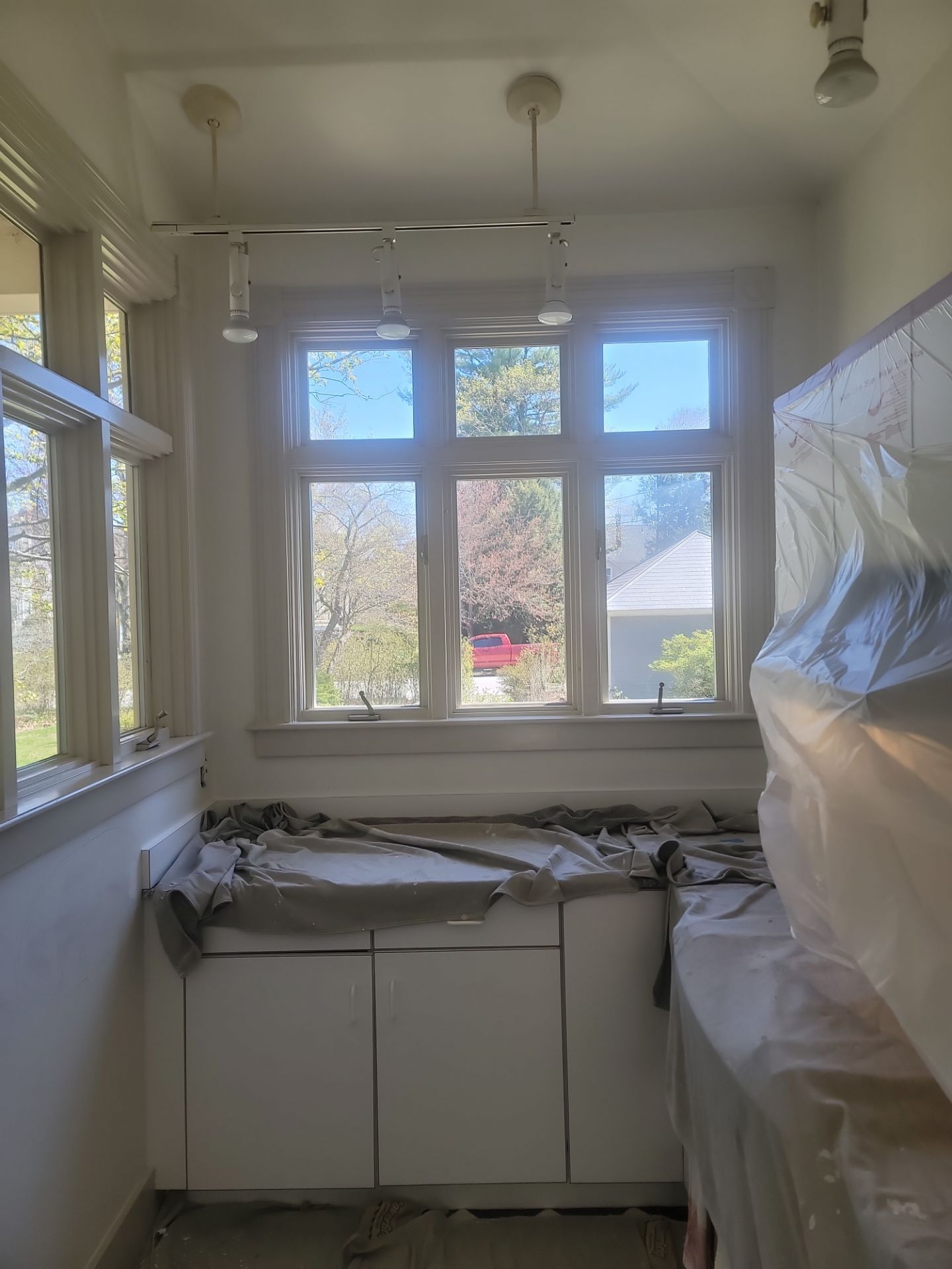 Small sunroom or kitchen under renovation, with covered cabinets, plastic sheeting, and bright windows.