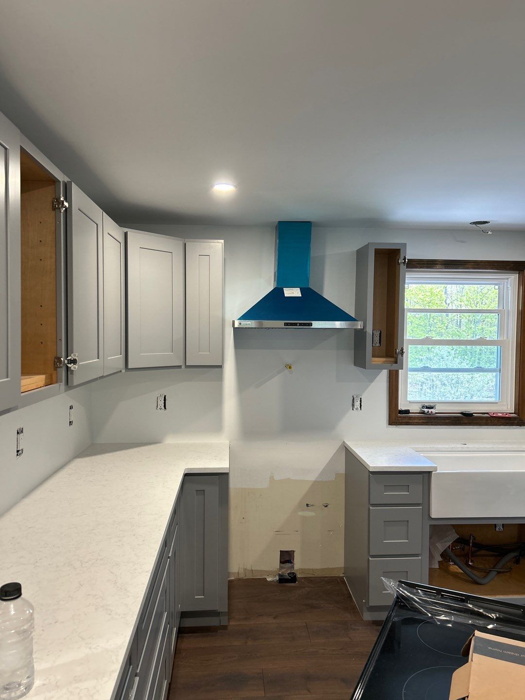Partially renovated kitchen with white cabinets, blue range hood, gray counters, and a window over a sink area