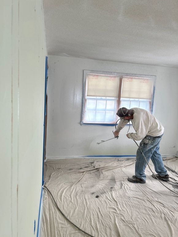Person painting a room white with a roller, standing on a covered floor near a window.