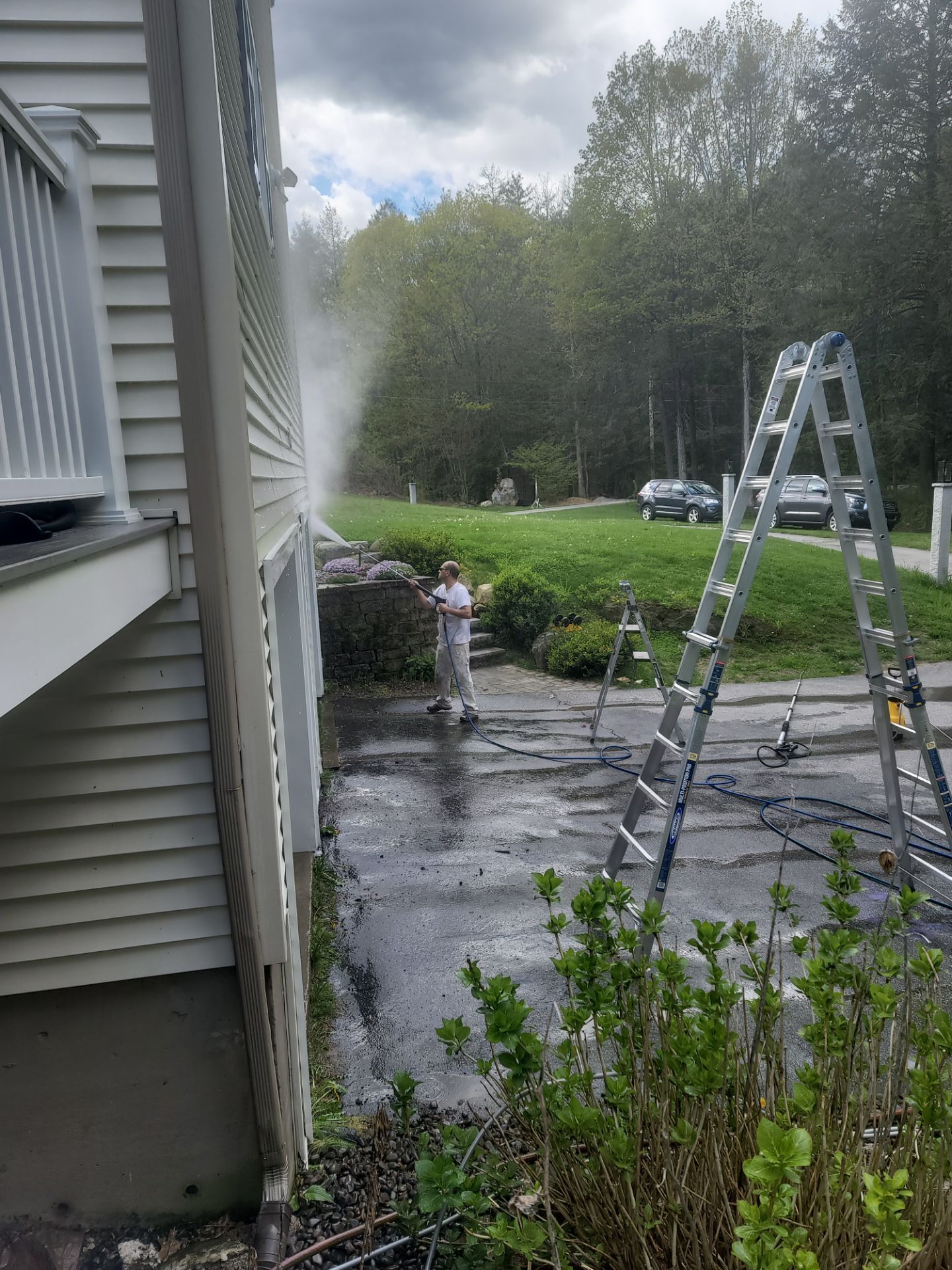 House exterior with ladder on wet driveway and water spraying from the left side of the building