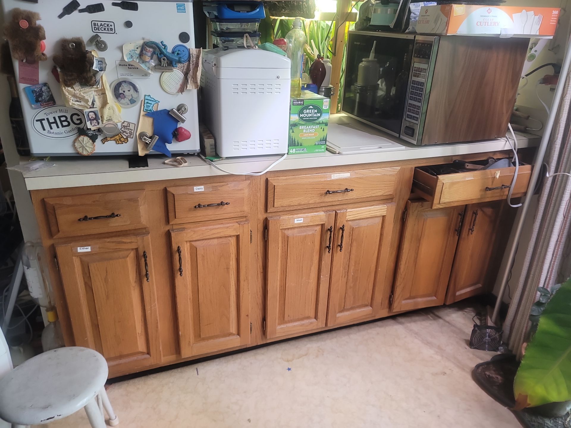 Wooden kitchen cabinets with cluttered countertop, microwave, bottles, and a small stool on the left.