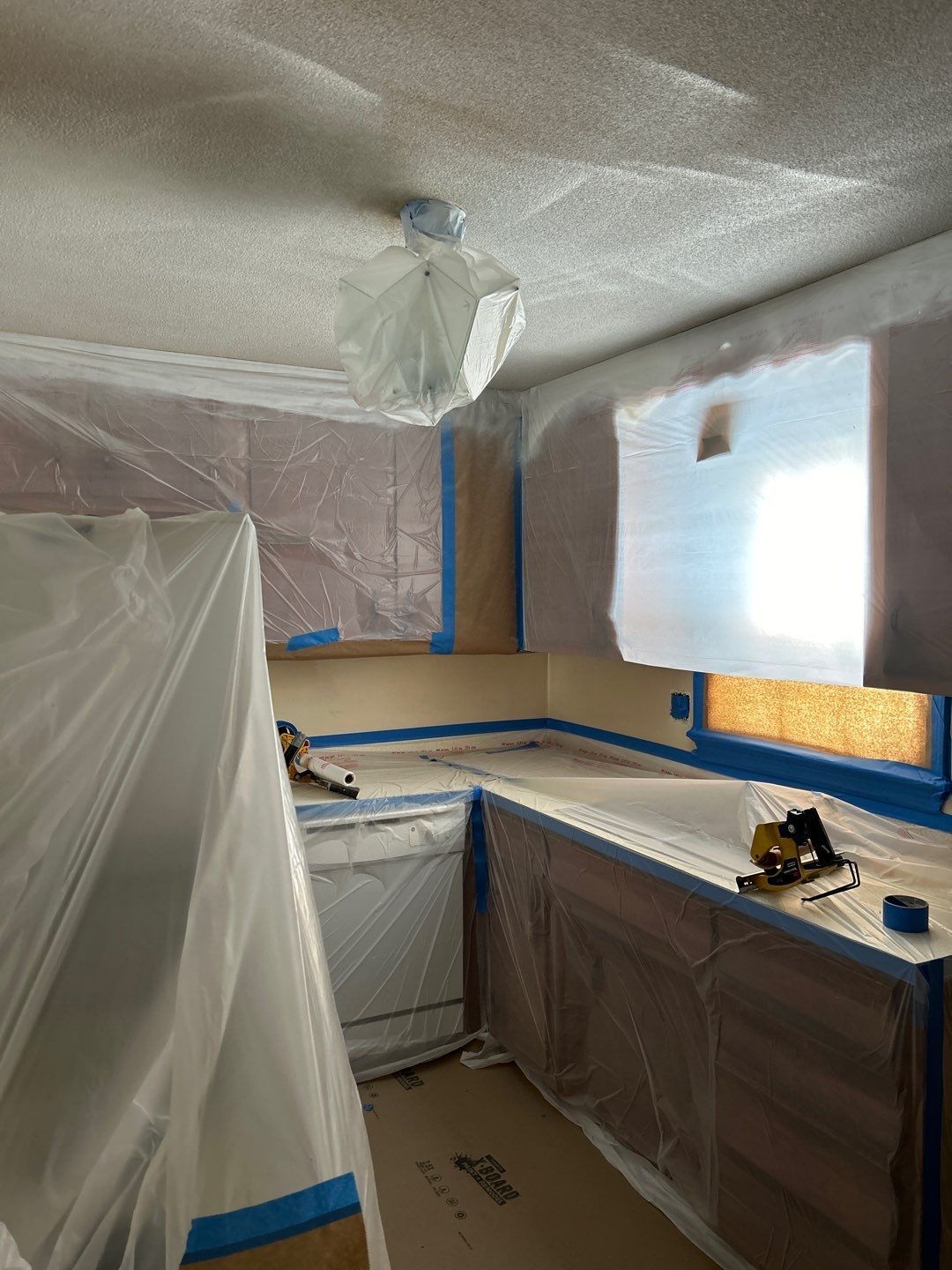 Plastic-covered kitchen under renovation with taped-off cabinets, countertop, and window.