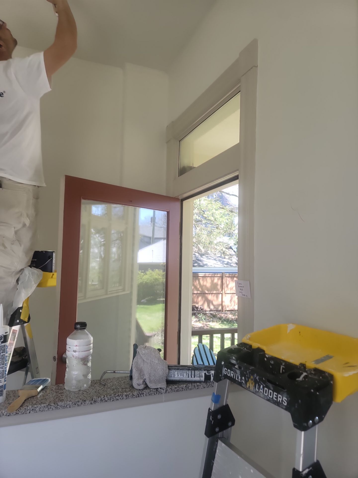 Person painting a room, with a red door, open window, and paint supplies on a counter.