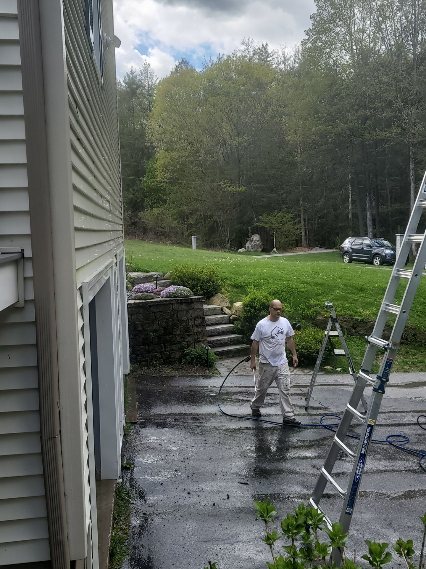 Worker pressure washing driveway beside house, with ladder and stone wall in the background.