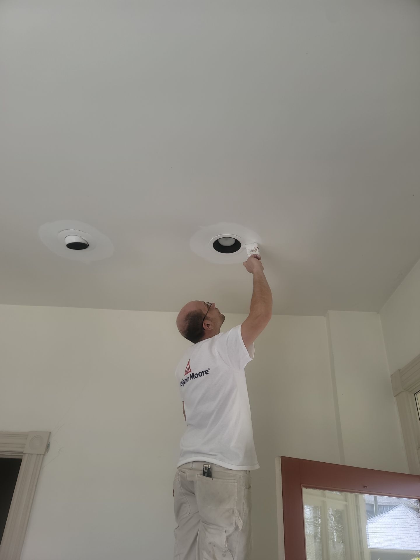 Person painting a white ceiling near two recessed lights in a room with a door and window