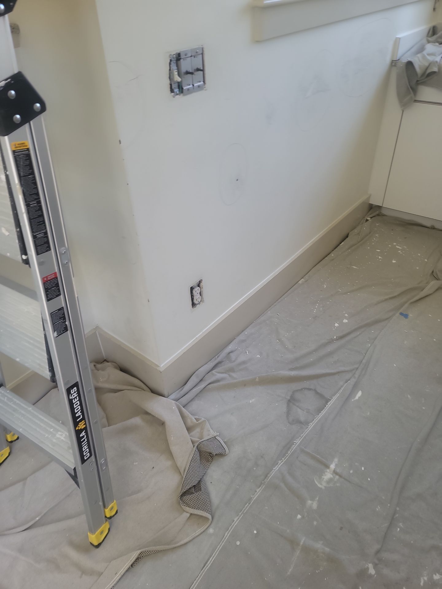 Painted wall and baseboard with plastic floor covering, ladder at left, in a room under repair
