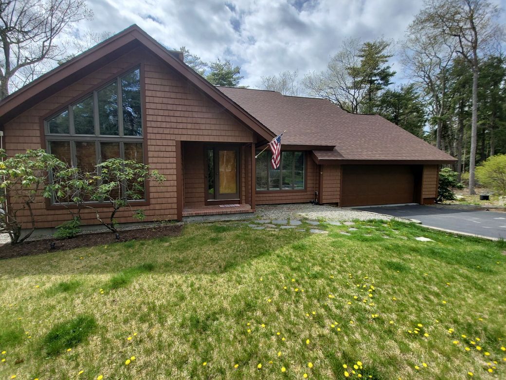 Brown house with large front window, garage, and American flag in a grassy yard under trees