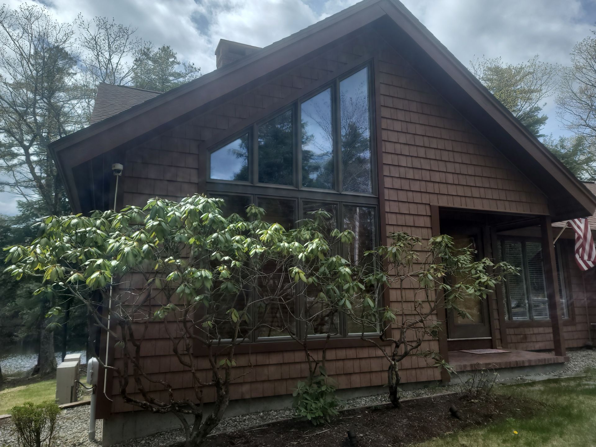 Brown cabin with large triangular windows, surrounded by shrubs and trees under a cloudy sky