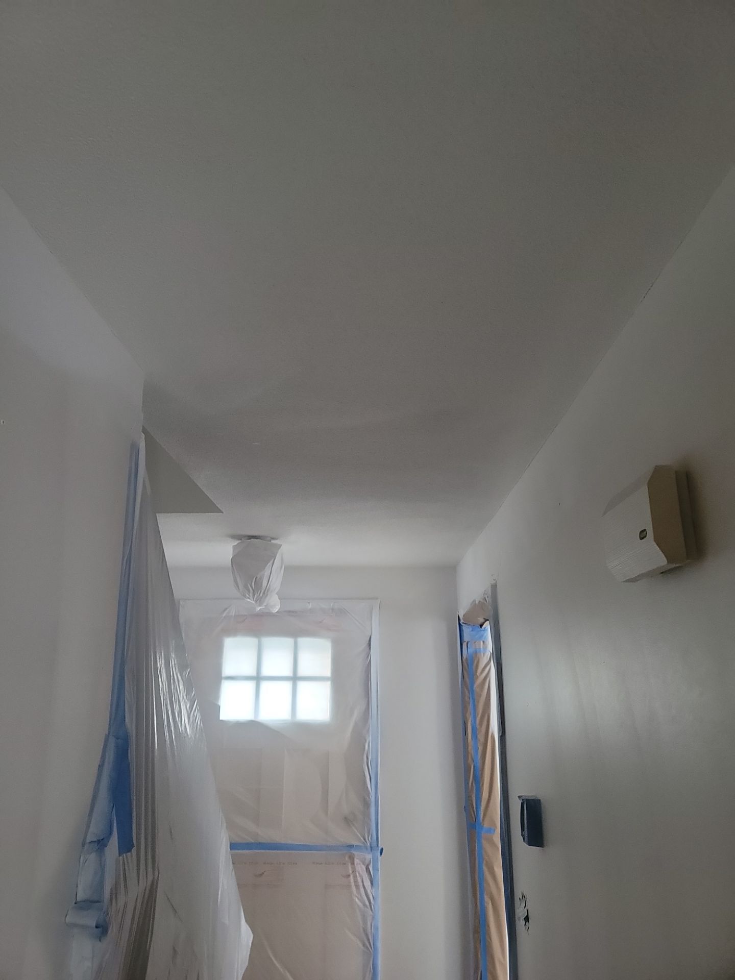 Narrow white hallway with a small window and blue tape covering parts of the walls and doorways