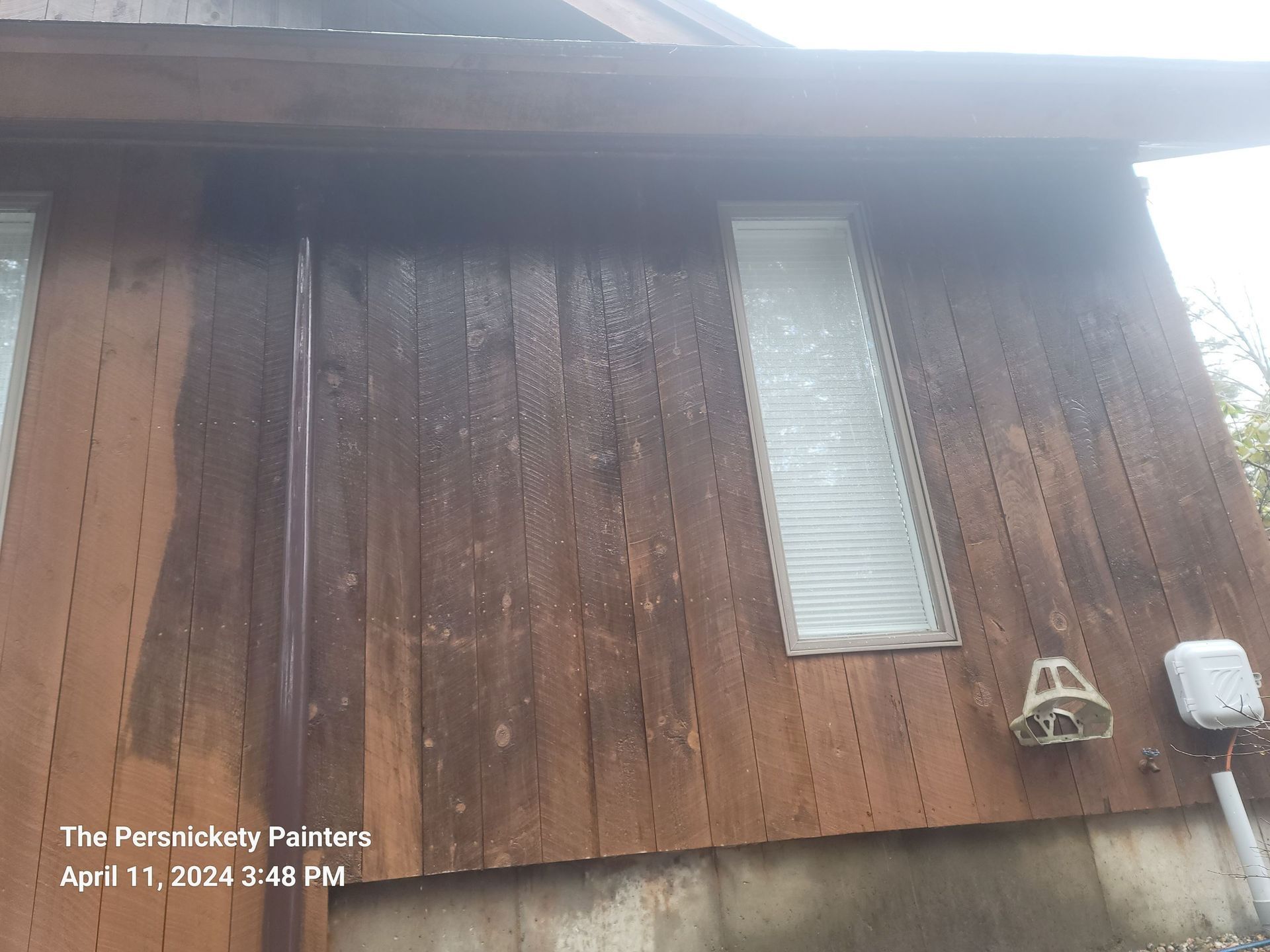 Exterior of a wooden house with wet vertical siding, a tall narrow window, and utility meters near the corner.