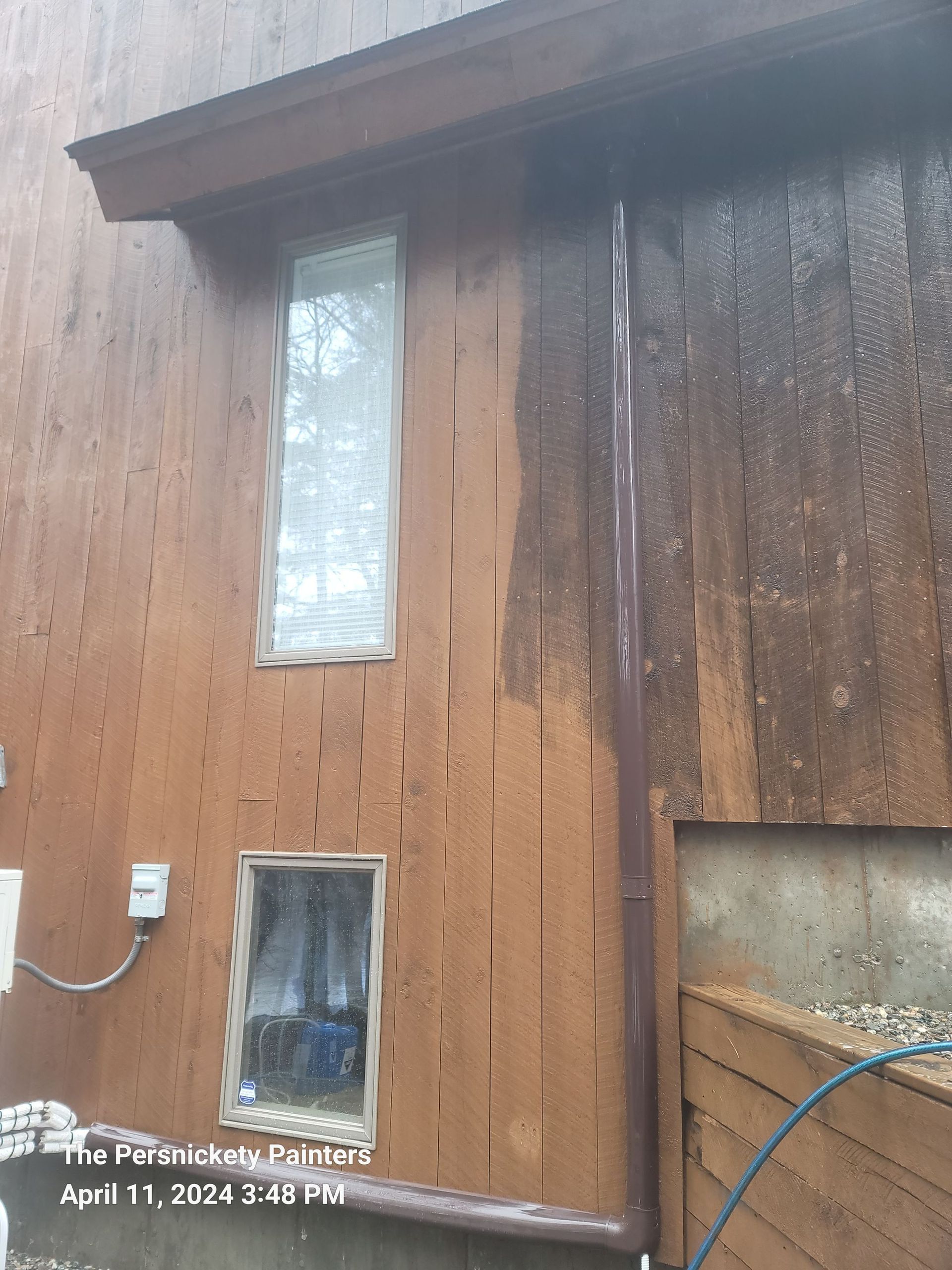 Weathered wooden house exterior with two windows and a blue hose along the right side.