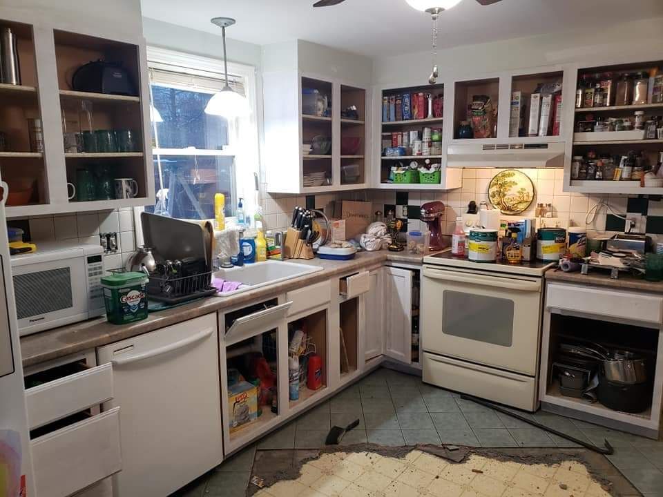 Cluttered kitchen with white cabinets, microwave, stove, shelves, and assorted items on counters and floor.