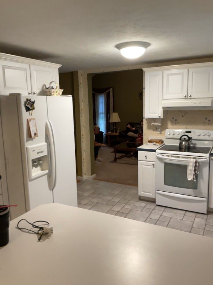 Kitchen with white cabinets, stove, and refrigerator, opening to a dim dining area.