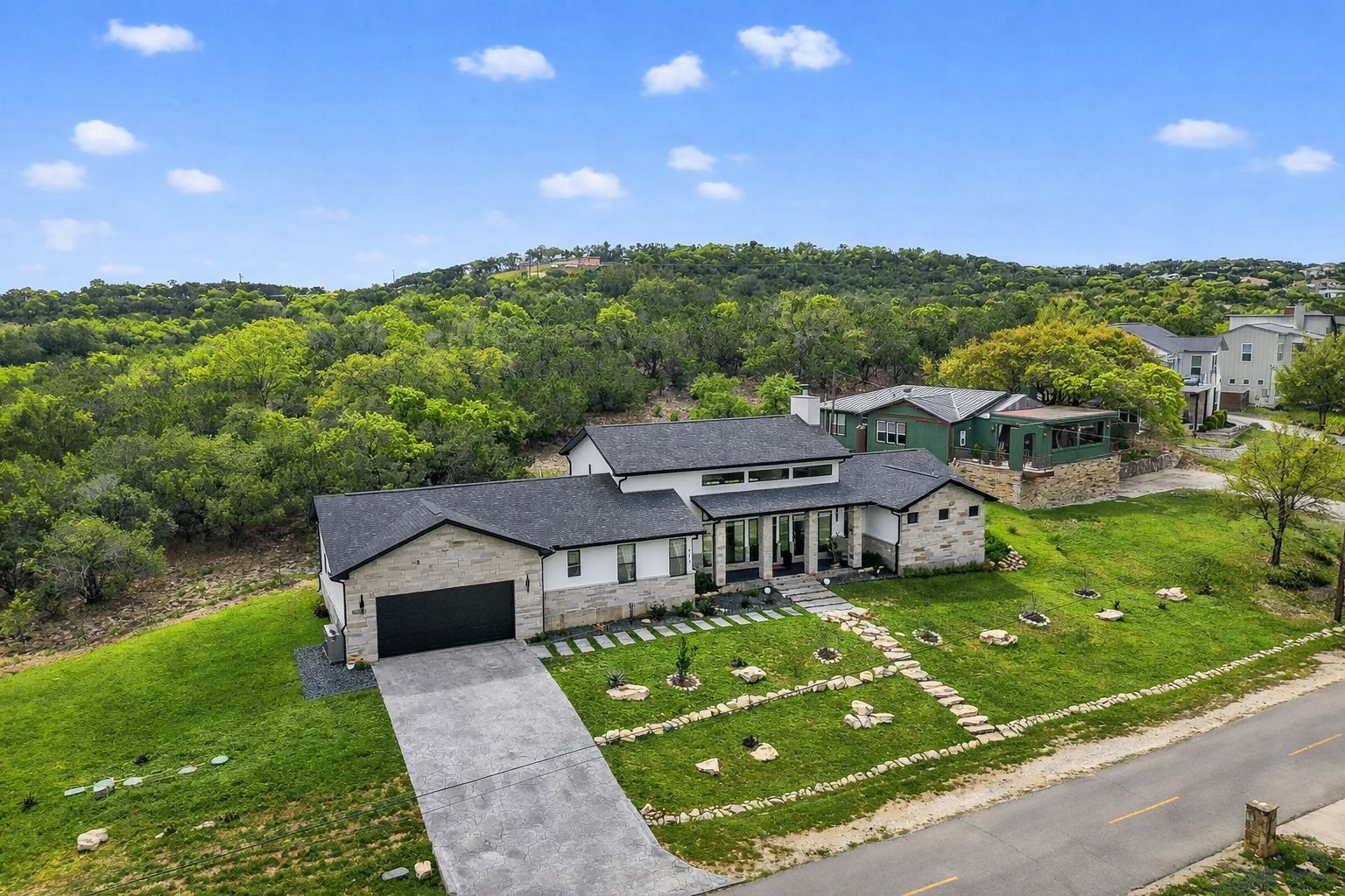 Aerial view of a large modern house with a gray roof, driveway, and green hills behind it.